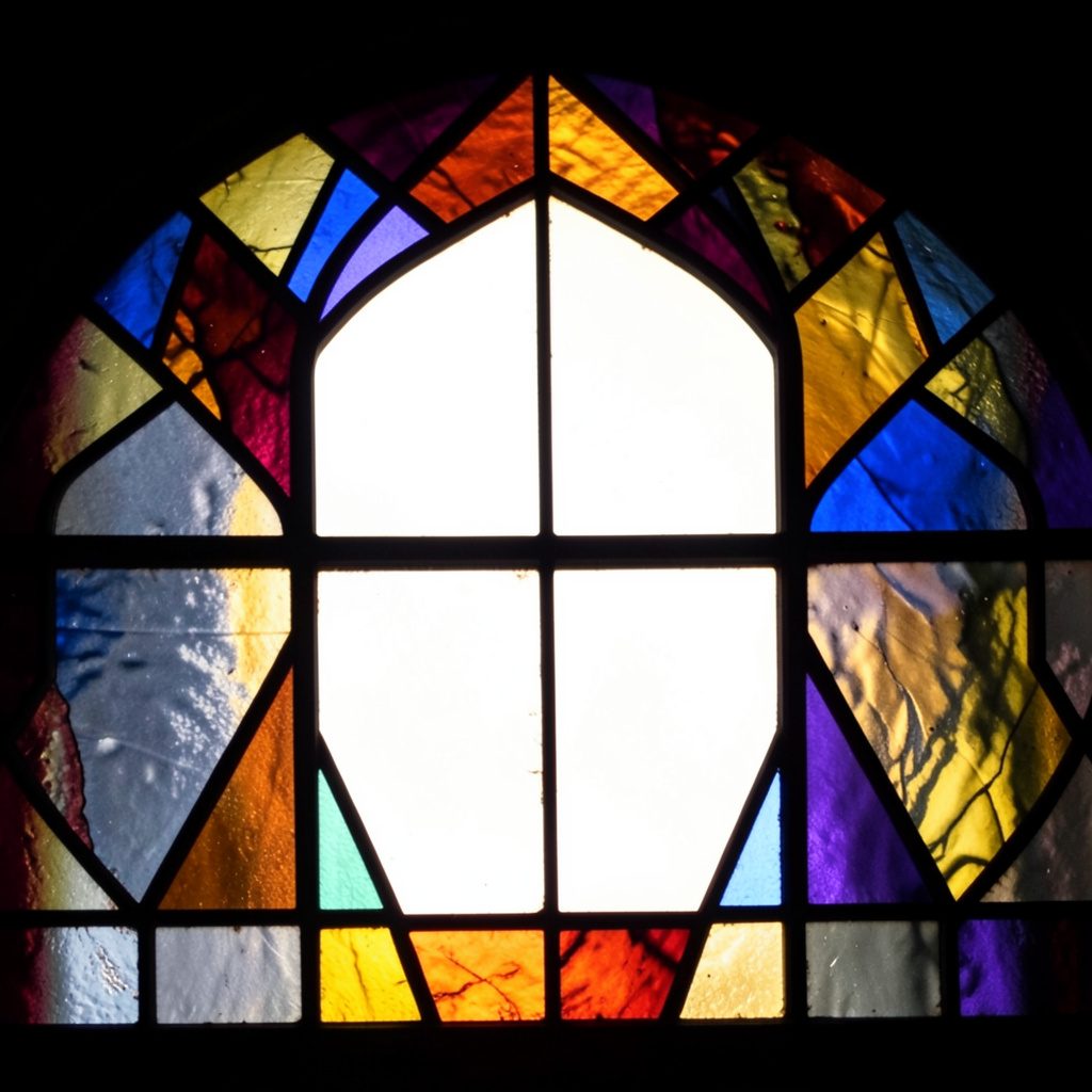 A dramatic stained-glass composition with bold leaded segments in deep ruby red, cobalt blue, and amber gold, glowing with transmitted light against a dark background.